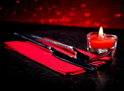 Valentine day table setting with knife, fork, red burning heart shaped candle on red hearts bokeh background