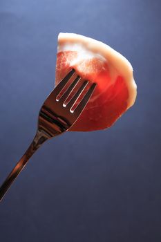 Italian prosciutto. Thin piece of pork meat on fork against dark background