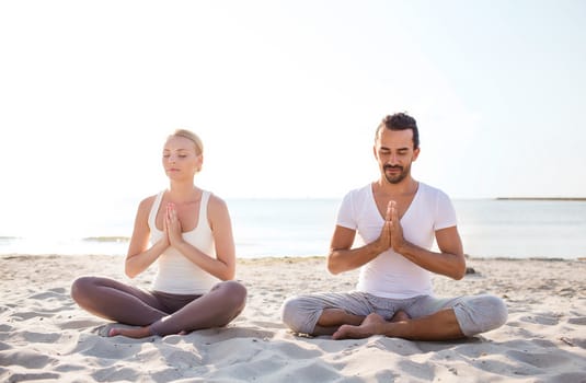 fitness, sport, friendship and lifestyle concept - smiling couple making yoga exercises sitting on sand outdoors