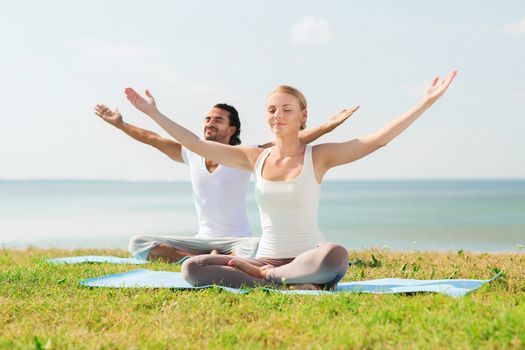 fitness, sport, friendship and lifestyle concept - smiling couple making yoga exercises sitting on mats outdoors