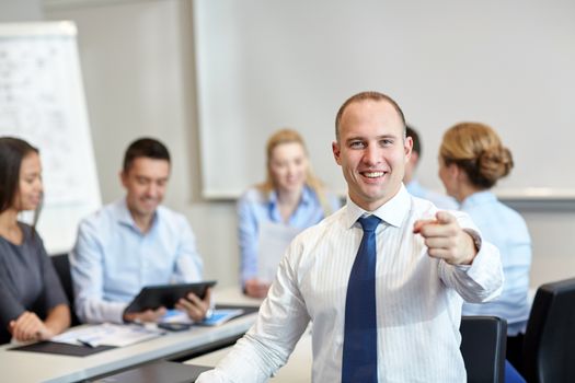 business, people, gesture and teamwork concept - smiling businessman pointing finger to you with group of businesspeople meeting in office