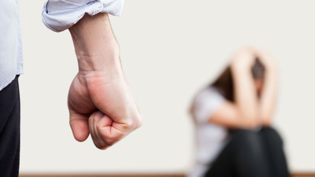 Family violence and aggression concept - furious angry man raised punishment fist over scared or terrified woman sitting at wall corner