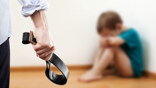 Family violence and aggression concept - furious angry man raised punishment hand holding leather belt over scared or terrified child boy sitting at wall corner