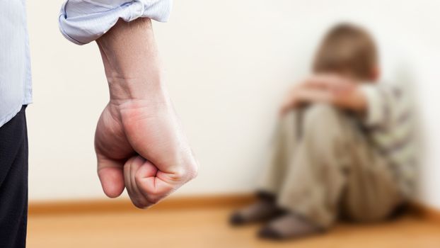 Family violence and aggression concept - furious angry man raised punishment fist over scared or terrified child boy sitting at wall corner