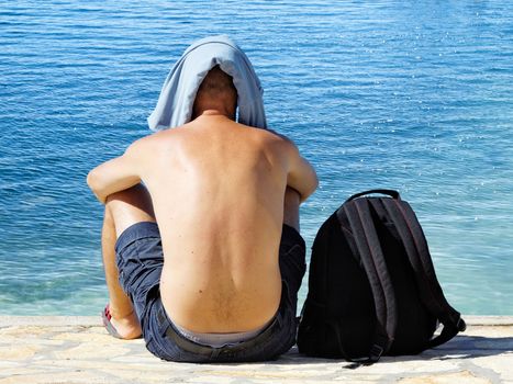 young man sitting on coastline with t-shirt on his head