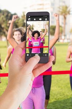 Female hand holding a smartphone against cheering young woman winning breast cancer marathon