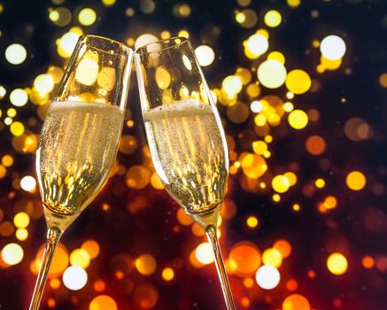 two champagne flutes with golden bubbles make cheers on colorful light bokeh background with space for text
