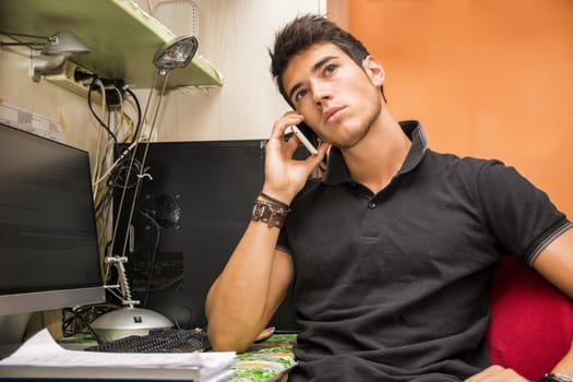 Waist Up Closeup of Young Attractive Man with Dark Hair Sitting at Computer Desk Talking on Cell Phone in Dorm Room