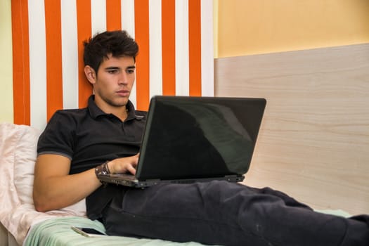 Attractive Young Man with Serious Expression, Reclining Comfortably with Laptop on Bed Working on his Start-up Business - Young Male College or University Student Doing Homework, in Bedroom