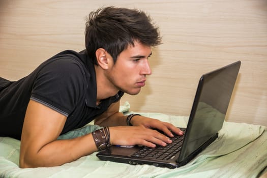 Attractive Young Man with Serious Expression, with Laptop on Bed Working on his Start-up Business - Young Male College or University Student Doing Homework, in Bedroom