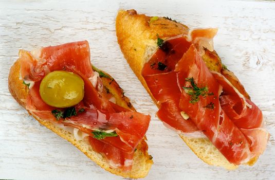 Delicious Tapas with Smoked Hamon on Garlic Bread with Green Olives and Olive Oil and closeup on Textured White Wooden background. Top View