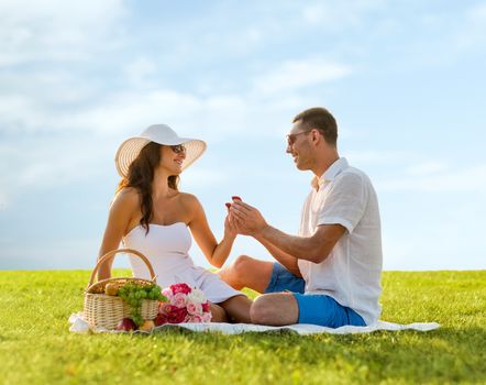 love, dating, people, proposal and holidays concept - smiling young man giving small red gift box with wedding ring to his girlfriend on picnic over blue sky and grass background