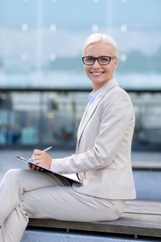 business, people and education concept - young smiling businesswoman in glasses with notepad over office building