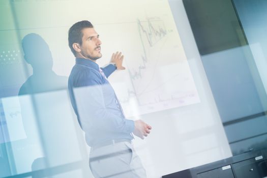 Business man making a presentation in front of whiteboard. Business executive delivering a presentation to his colleagues during meeting or in-house business training. View through glass.