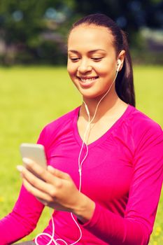 fitness, park, technology and sport concept - smiling african american woman with smartphone and earphones sitting on mat outdoors