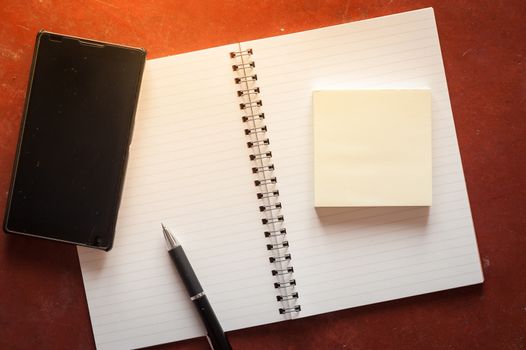 Opened notepad with pen, small sticky paper and black smart phone on rustic cement background with low key scene.