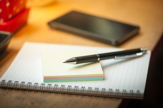 Opened notepad with pen, small sticky paper and black smart phone on wood bar in coffee shop in morning scene