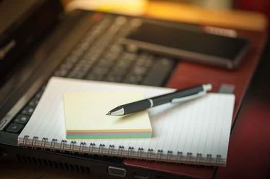 Opened notepad with pen, small sticky paper and black smart phone on laptop computer in coffee shop with morning scene