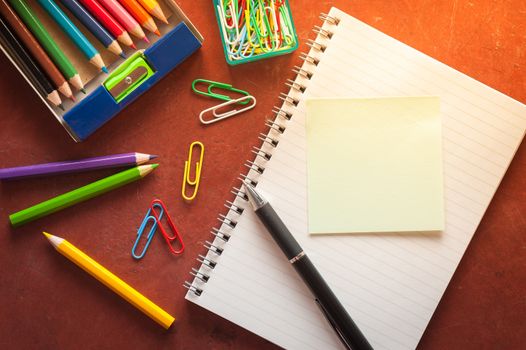 Opened notepad with pen, small sticky paper, colored pencils, and paper clips on red cement background with morning scene