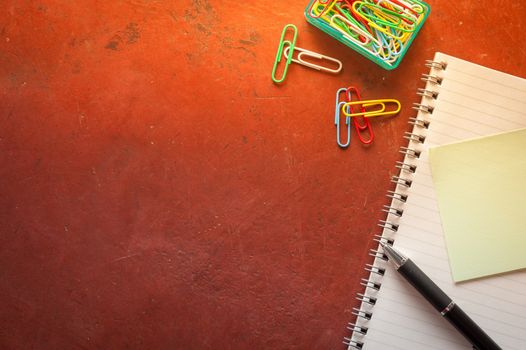 Opened notepad with pen, small sticky paper, and paper clips on red cement background with morning scene