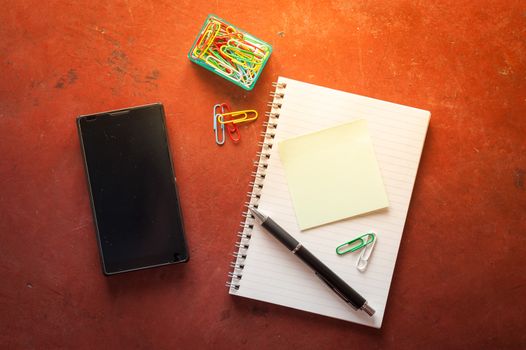 Opened notepad with pen, small sticky paper, smartphone, and paper clips on red cement background with morning scene