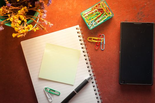 Opened notepad with pen, small sticky paper, smartphone, and paper clips on red cement background with morning scene