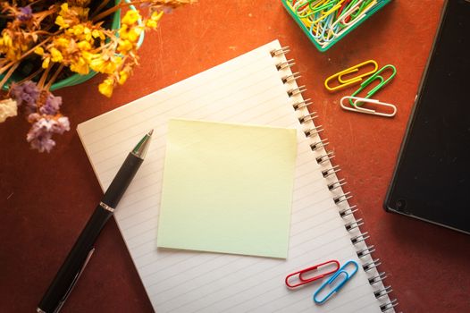 Opened notepad with pen, small sticky paper, smartphone, and paper clips on red cement background with morning scene