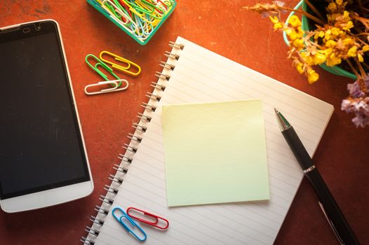 Opened notepad with pen, small sticky paper, smartphone, and paper clips on red cement background with morning scene