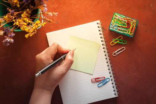 Opened notepad with pen, small sticky paper, and paper clips with female left hand writing on blank area on red cement background with morning scene