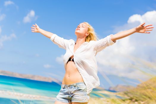 Relaxed woman enjoying freedom and life an a beautiful sandy beach.  Young lady raising arms, feeling free, relaxed and happy. Concept of freedom, happiness, enjoyment and well being.