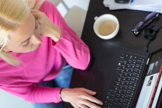 Female freelancer in her casual home clothing remotly working on laptop computer from her home while drinking her morning cup coffee. View from above.