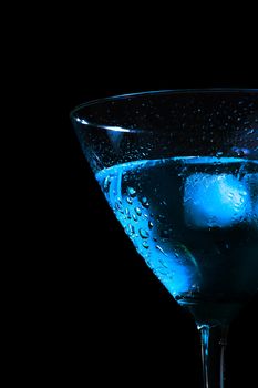 detail of half glass of fresh blue cocktail with ice on blue tint light and black background with space for text