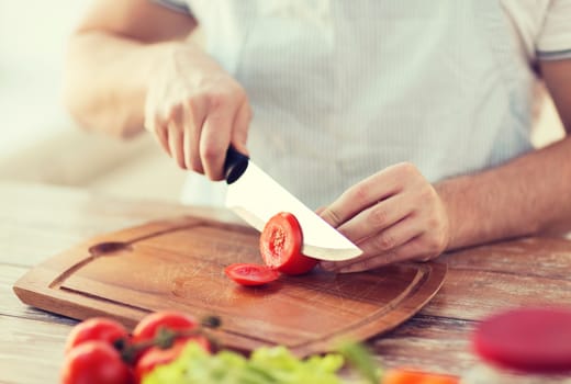 cooking and home concept - close up of male hand cutting tomato on cutting board with sharp knife