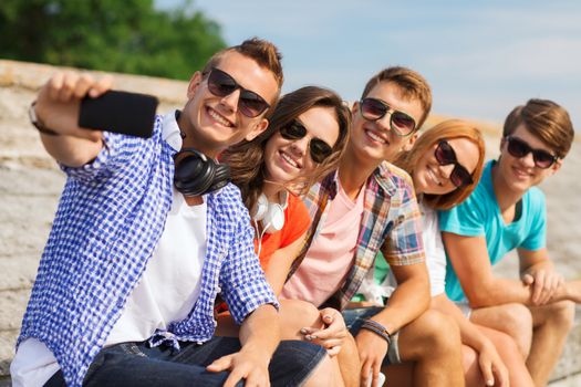 friendship, summer, technology and people concept - group of smiling friends with smartphone and headphones making selfie outdoors