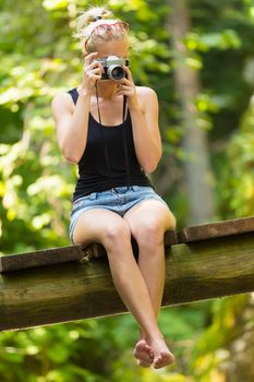 Beautiful barefooted blonde caucasian girl wearing jeans shorts an sporty black sleeveless t-shirt, sitting on a vintage wooden bridge, taking photos with retro camera.