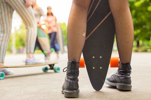 Teenagers practicing long board riding outdoors in skateboarding park. Detail of  legs of girl wearing black boots and stockings holding long board in foreground. Active urban life. Urban subculture.