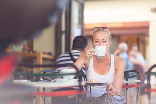 Calm casual blond lady enjoying cup of coffee outdoor in typical italian  street coffee house on warm summer day.