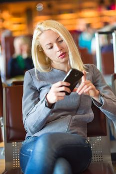 Casual blond young woman using her cell phone while waiting to board a plane at the departure gates. Wireless network hotspot enabling people to access internet conection. Public transport.