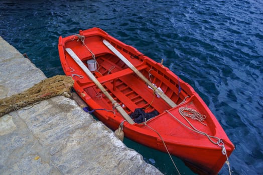 A beautiful red dory floats on the blue Aegean while tied to a grey granite pier.