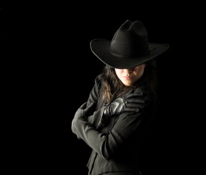 Brunette woman in black dress and black gloves, wearing a black cowboy hat, holding her arms across her body and looking downward, with her face partially hidden by the hat.