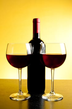 two red wine glasses near bottle on golden background and wood table