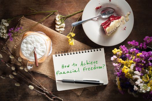"How to achieve financial freedom?" is written on Notepad with pen, a glass of Thai tea and bits of red velvet cake on rustic wood table with low key scene