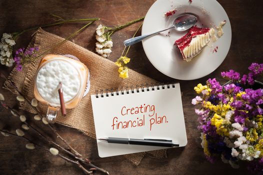 "Creating financial plan" is written on Notepad with pen, a glass of Thai tea and bits of red velvet cake on rustic wood background with low key scene