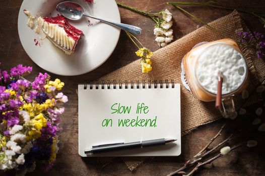 "Slow life on weekend" is written on Notepad with pen, a glass of Thai tea and bits of red velvet cake on rustic wood background with low key scene