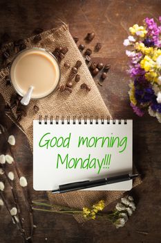 "Good morning monday" is written on Notepad with pen, a glass of Thai tea and bits of red velvet cake on rustic wood background with low key scene