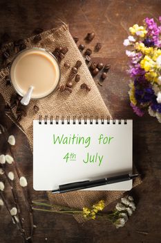 "waiting for 4th july" is written on Notepad with pen, a glass of Thai tea and bits of red velvet cake on rustic wood background with low key scene