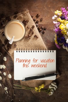 "Planning for vacation this weekend" is written on Notepad with pen, a glass of Thai tea and bits of red velvet cake on rustic wood background with low key scene