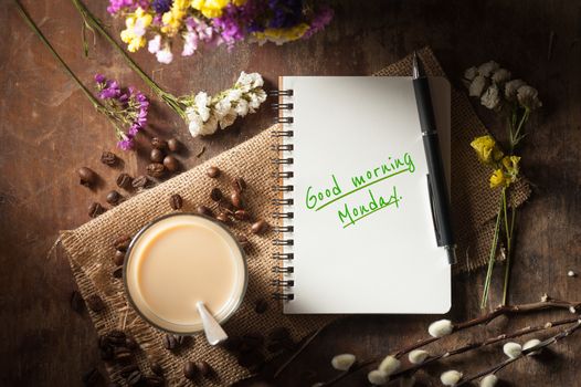 "Good morning monday" is written on Notepad with pen, a glass of Thai tea and bits of red velvet cake on rustic wood background with low key scene