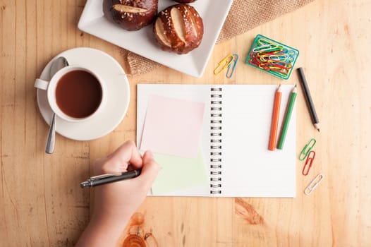 Notebook and sticky paper, paper clip, color pencil, and pen on wood table in vintage style with female left hand wrting on notepad