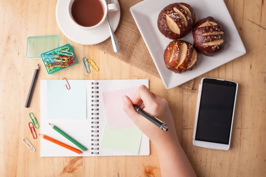 Notebook and sticky paper, paper clip, color pencil, smartphone, and pen on wood table in vintage style with female hand wrting on notepad